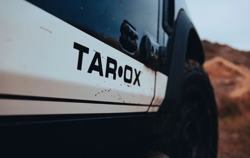 Close-up of a vehicle with the label 'TAROX' visible on the side, parked outdoors with a rocky landscape in the background.
