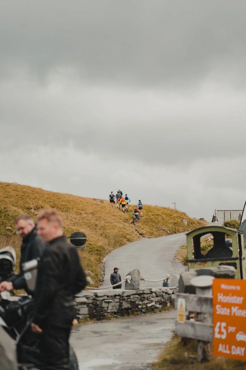 A group of hikers walking up a grassy hill on a cloudy day, with some blurred people and a sign in the foreground.