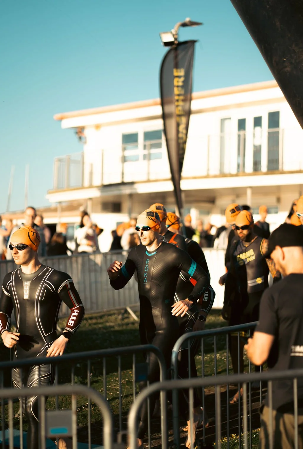Triathletes in wetsuits and orange swim caps preparing for a race near the water, with spectators and a building in the background.