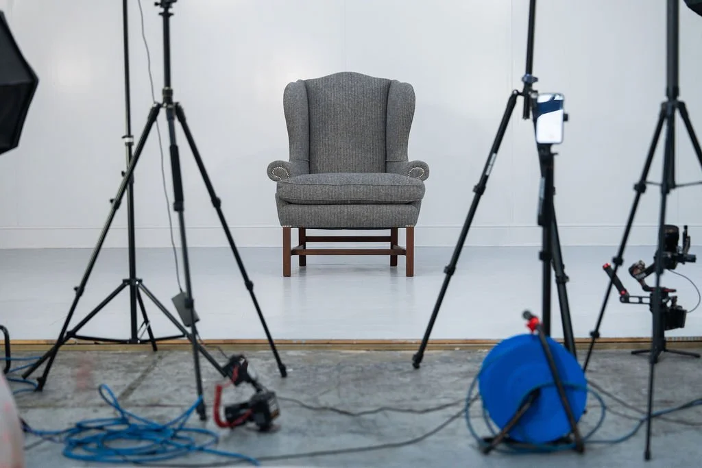 A gray upholstered armchair on a white studio floor with professional photography lighting and equipment surrounding it.