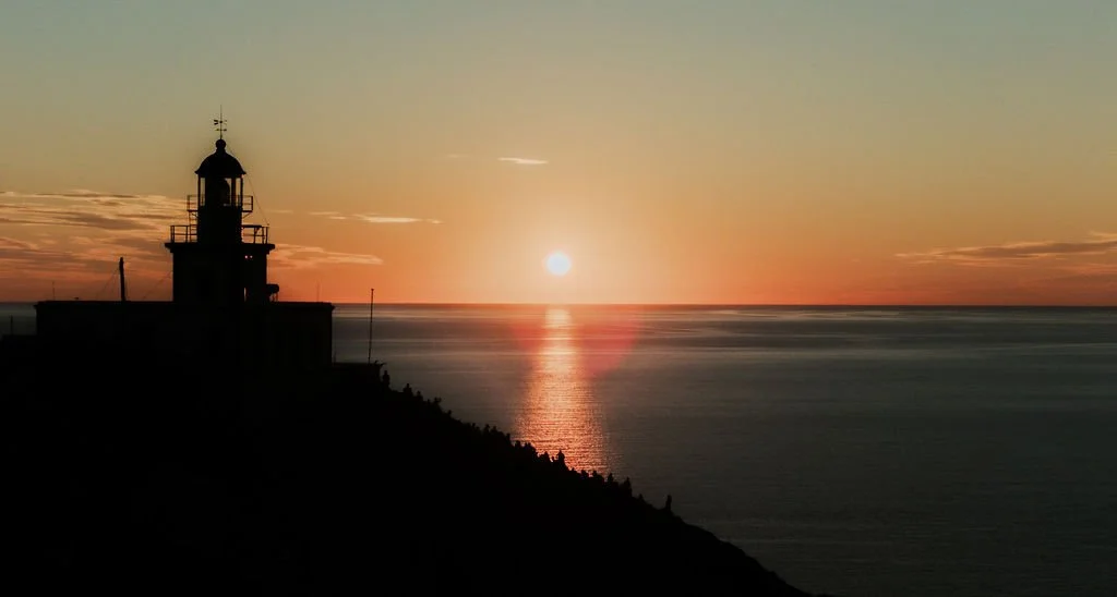 Sunset over the ocean with a lighthouse silhouette on the left.