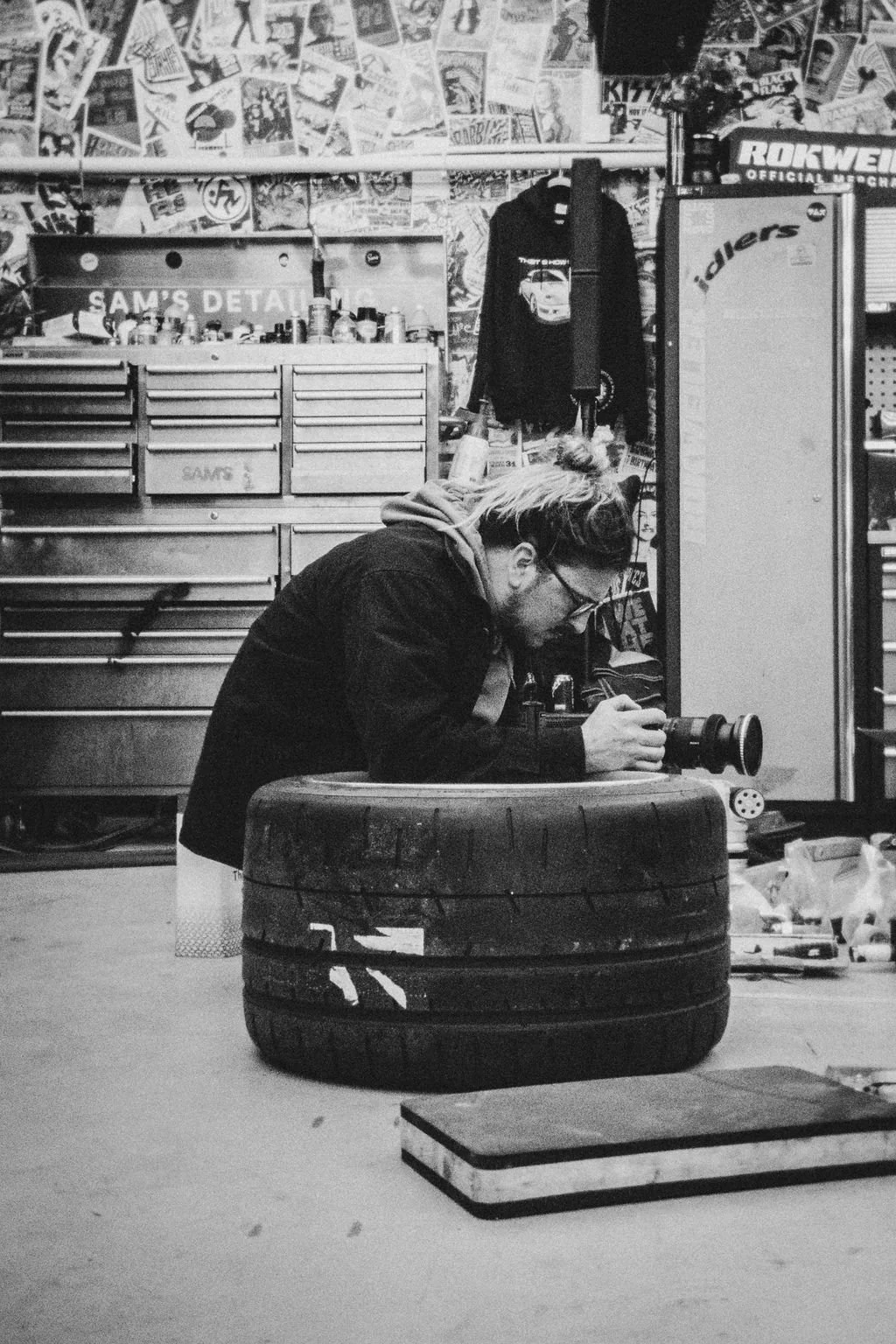 A man with glasses and blonde hair tied back, wearing a dark hoodie, is taking a photograph of a car tire with a camera, inside a garage or workshop cluttered with tools, posters, and locker storage.