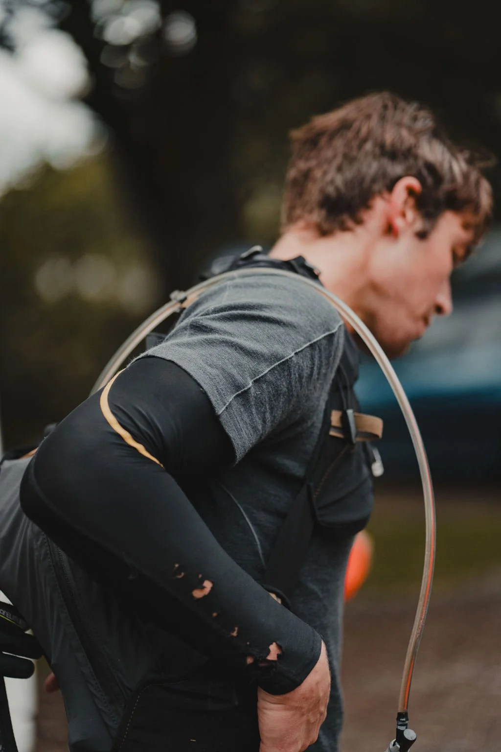 A person wearing a black and grey jacket with holes in the sleeve, bending over outside with trees in the background.