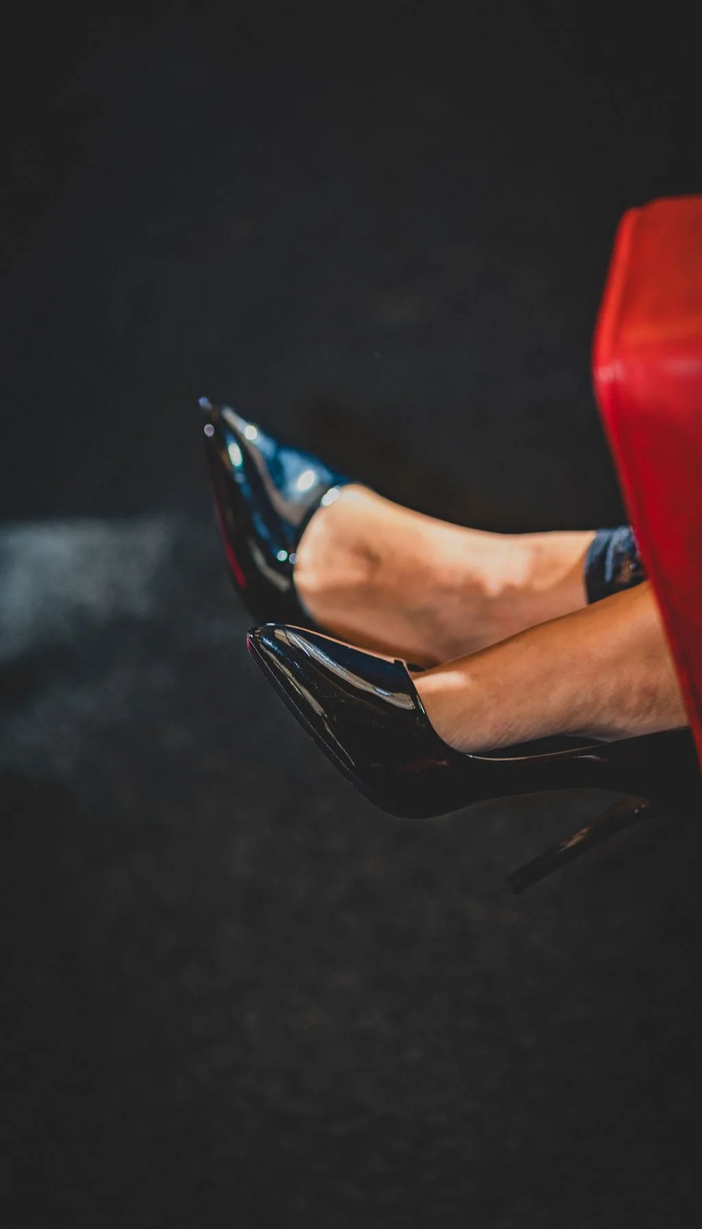 Close-up of a woman's legs wearing black high heels with a shiny finish, sitting on a red chair with a black background.