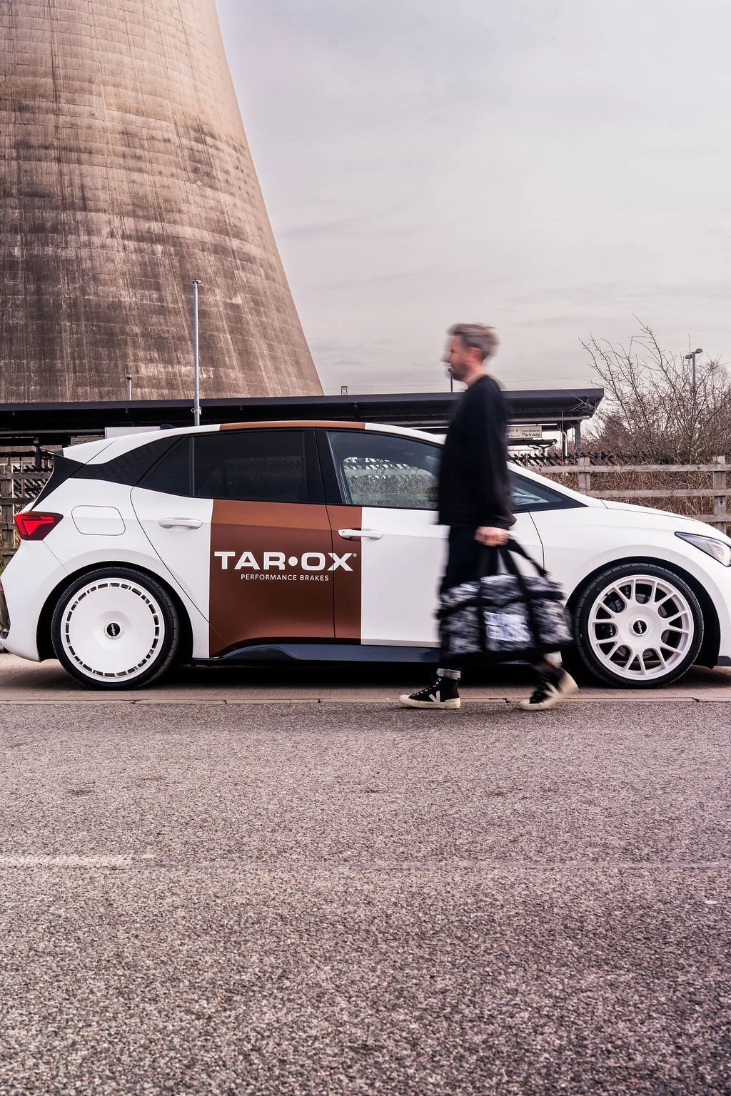 A man walking past a white car with branding for Tar-Ox performance brakes, carrying a black and white patterned bag.