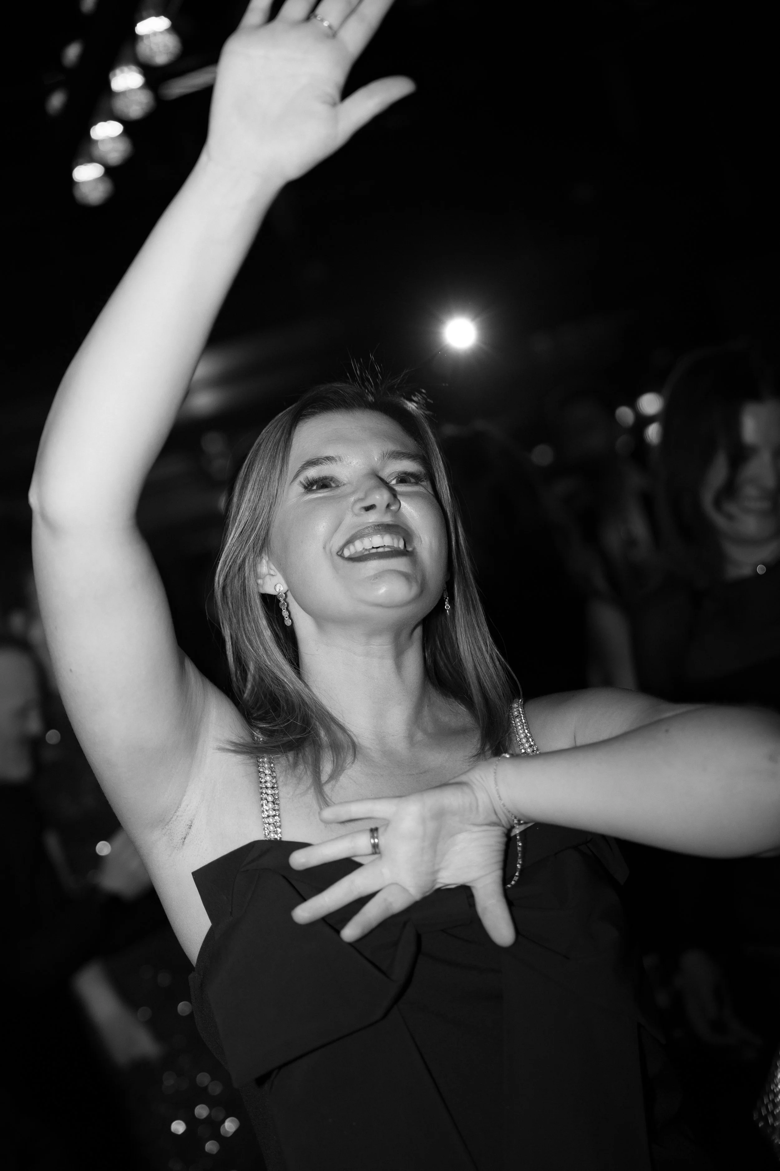 A woman smiling and dancing with her hand raised in a dark room, possibly at a party or celebration, black and white photograph.