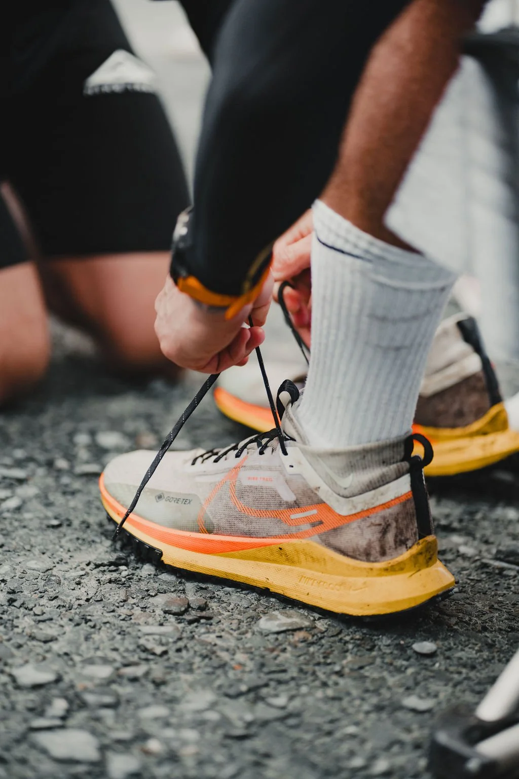 Person tying shoelaces on a running shoe on a gravel surface.