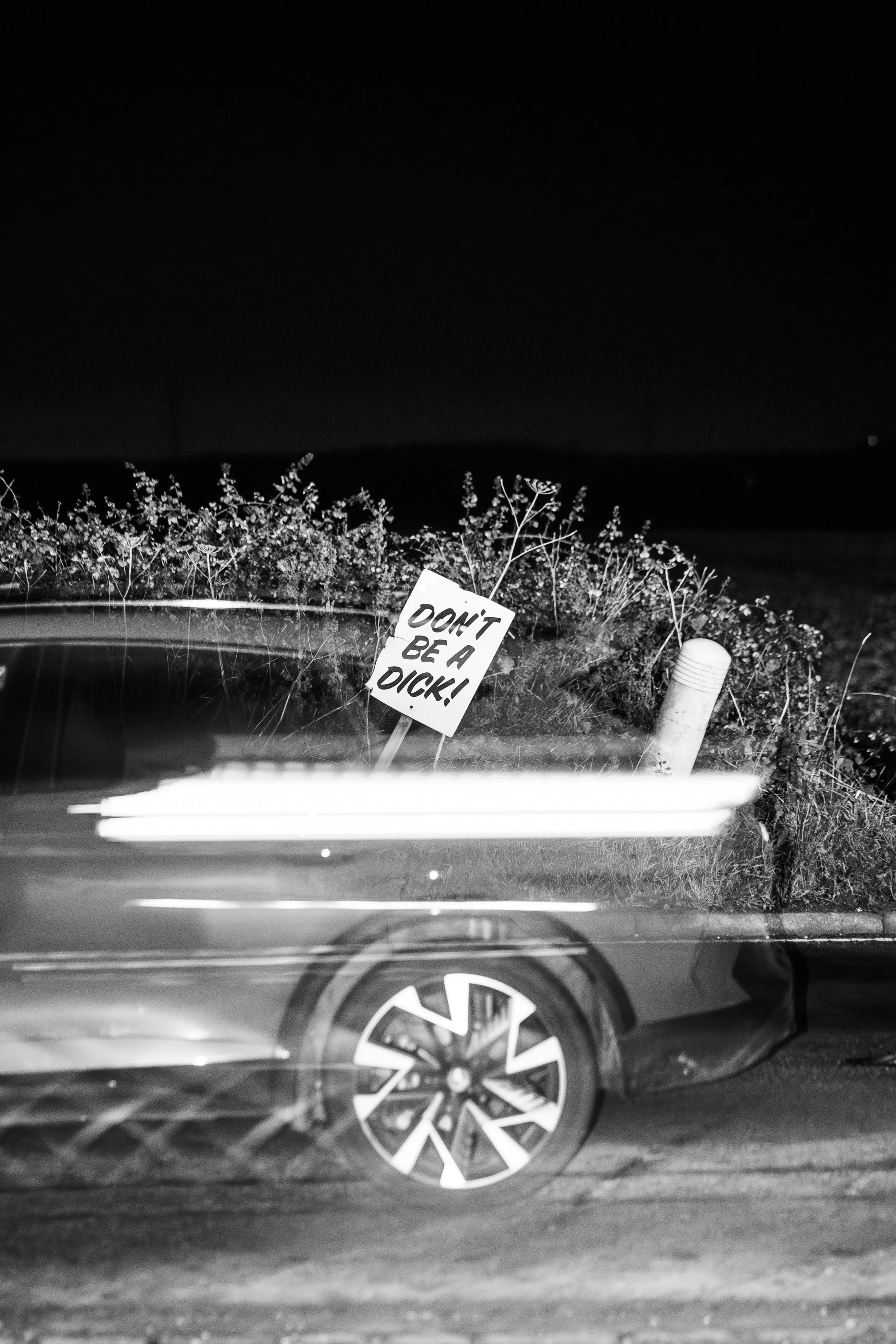 A black and white photo of a car parked on the side of a road at night. A sign in the background reads 'DON'T BE A DICK!' and there are some plants and a white pole nearby.