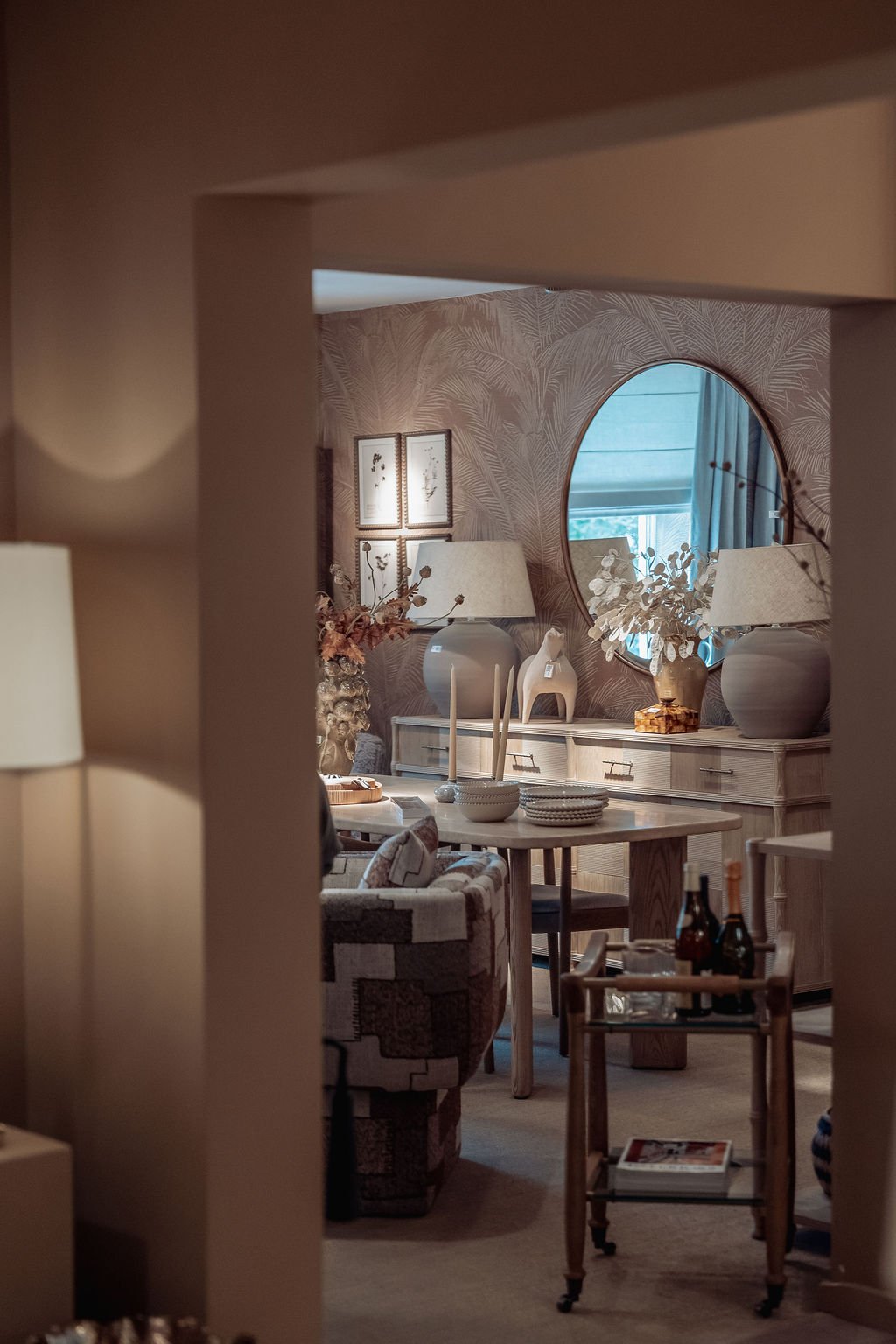 Decorative showroom with beige walls, a round mirror, two table lamps, and various vases with plants on a sideboard, viewed through a doorway.