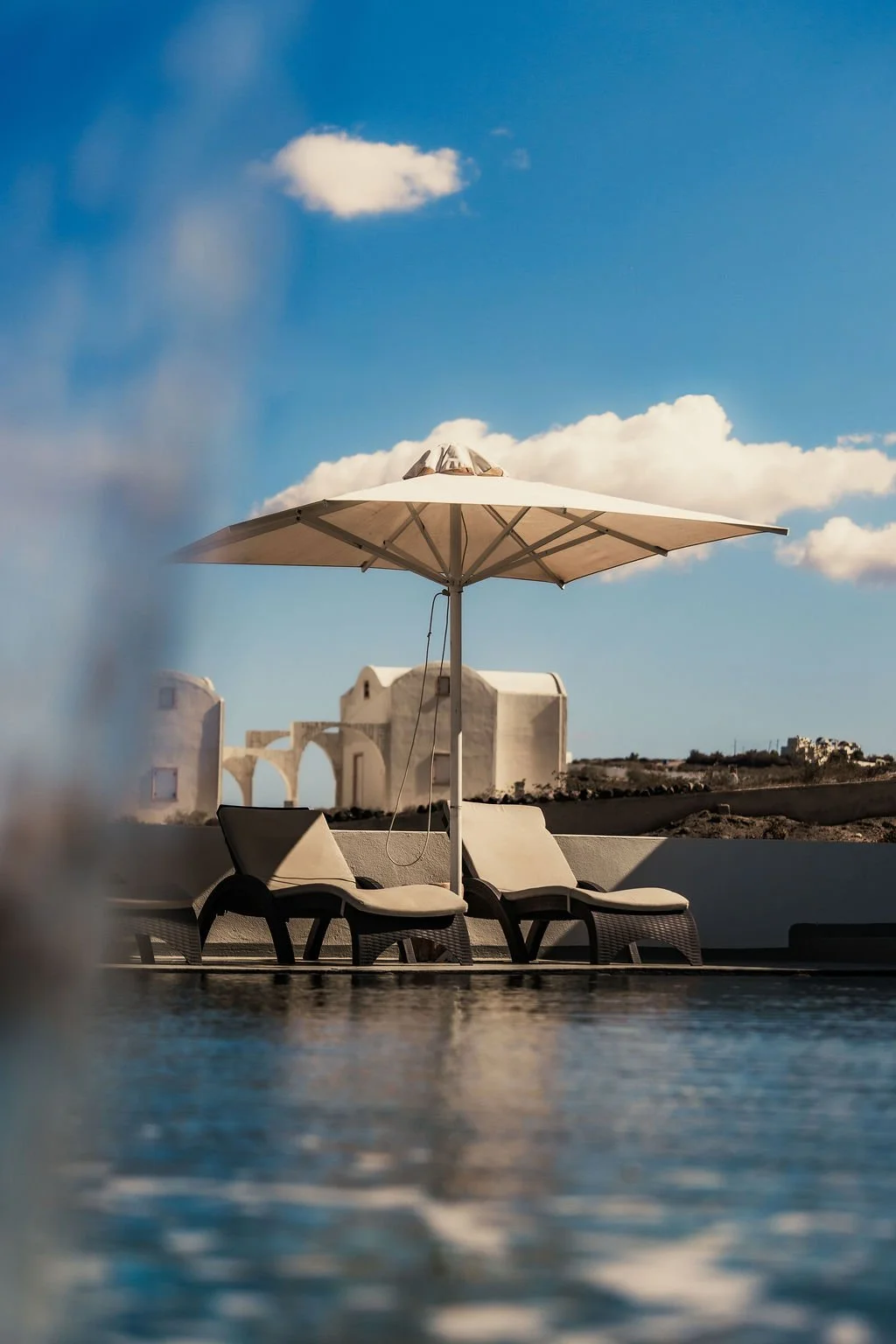 Two lounge chairs with cushions and a large umbrella by a swimming pool with a white building and cloudy sky in the background.