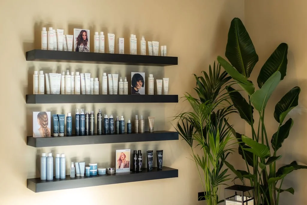 Display of hair care products on black shelves next to large green plants in a salon or spa.