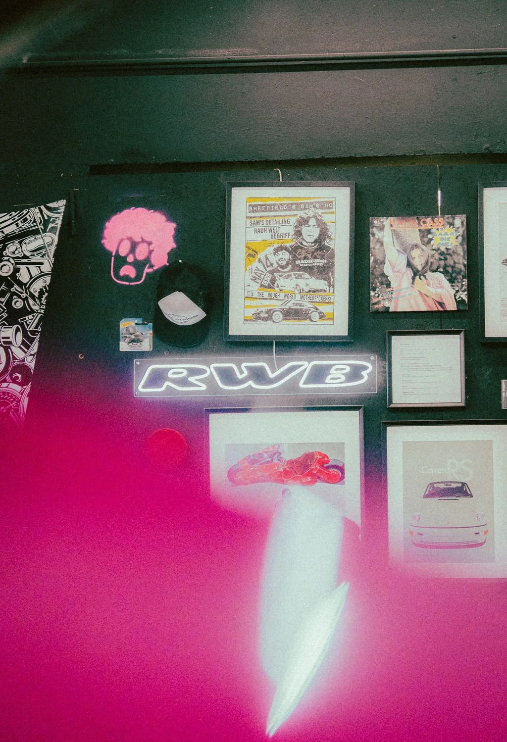 Wall decorated with various framed pictures and posters, a neon sign reading 'RWB', a pink skull illustration with curly hair, a baseball cap hanging on the wall, and pink and white lighting effects.