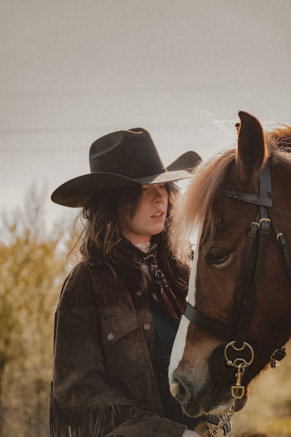 Young woman with a black cowboy hat, brown jacket, and bandana, standing next to a brown and white horse outdoors, with trees and cloudy sky in the background.
