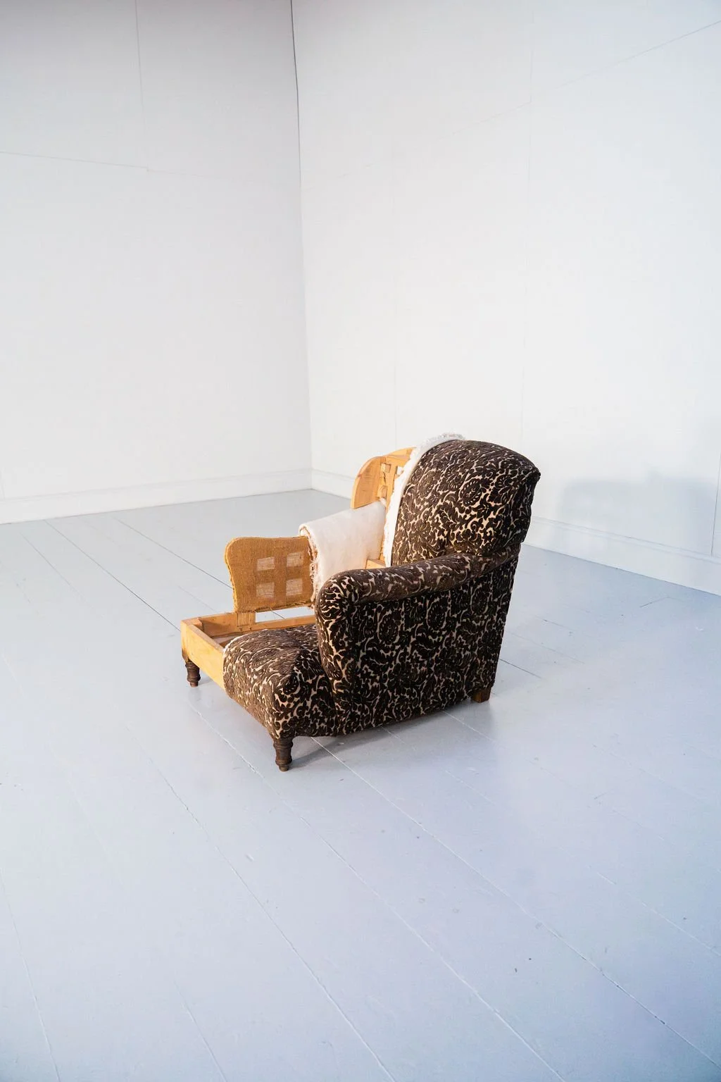 A vintage armchair with black and gold floral upholstery, placed against a white wall and floor, with parts of a wooden frame and furniture padding visible.