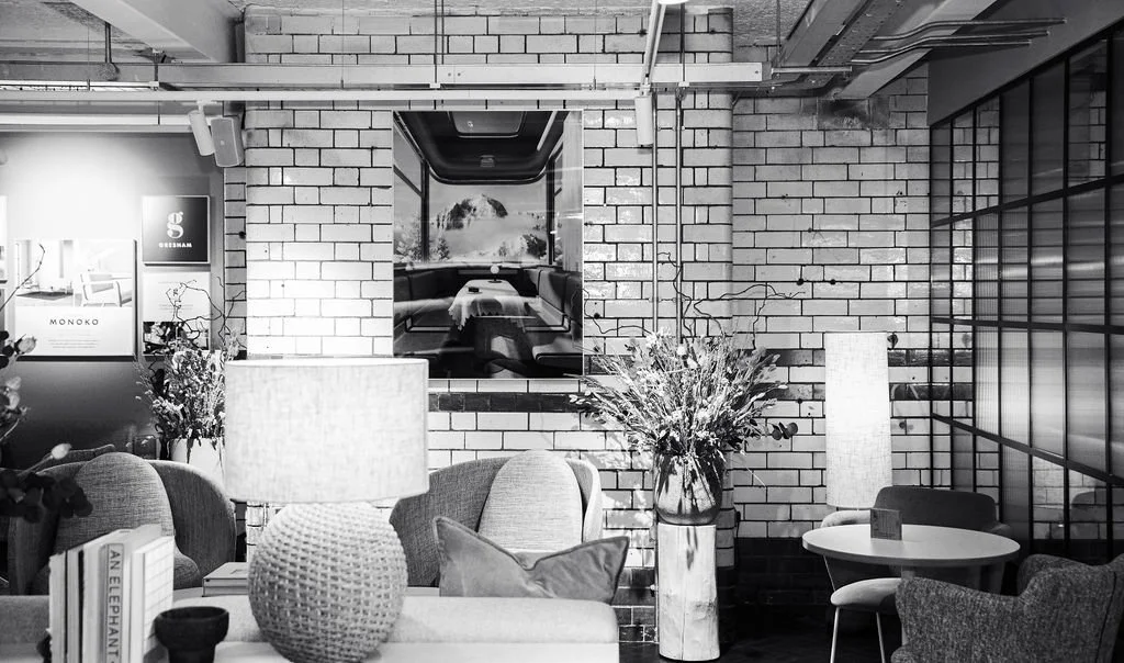 Interior of a modern office space with white brick walls, plants, lamps, and furniture in black, white, and grey tones.