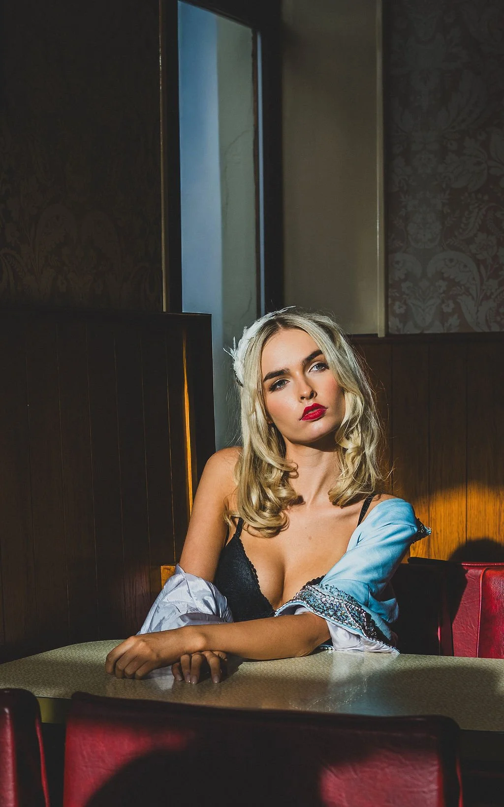A woman with blonde hair and red lipstick sitting at a table in a dimly lit restaurant or bar, wearing a black bra and a partially open blue satin shirt.