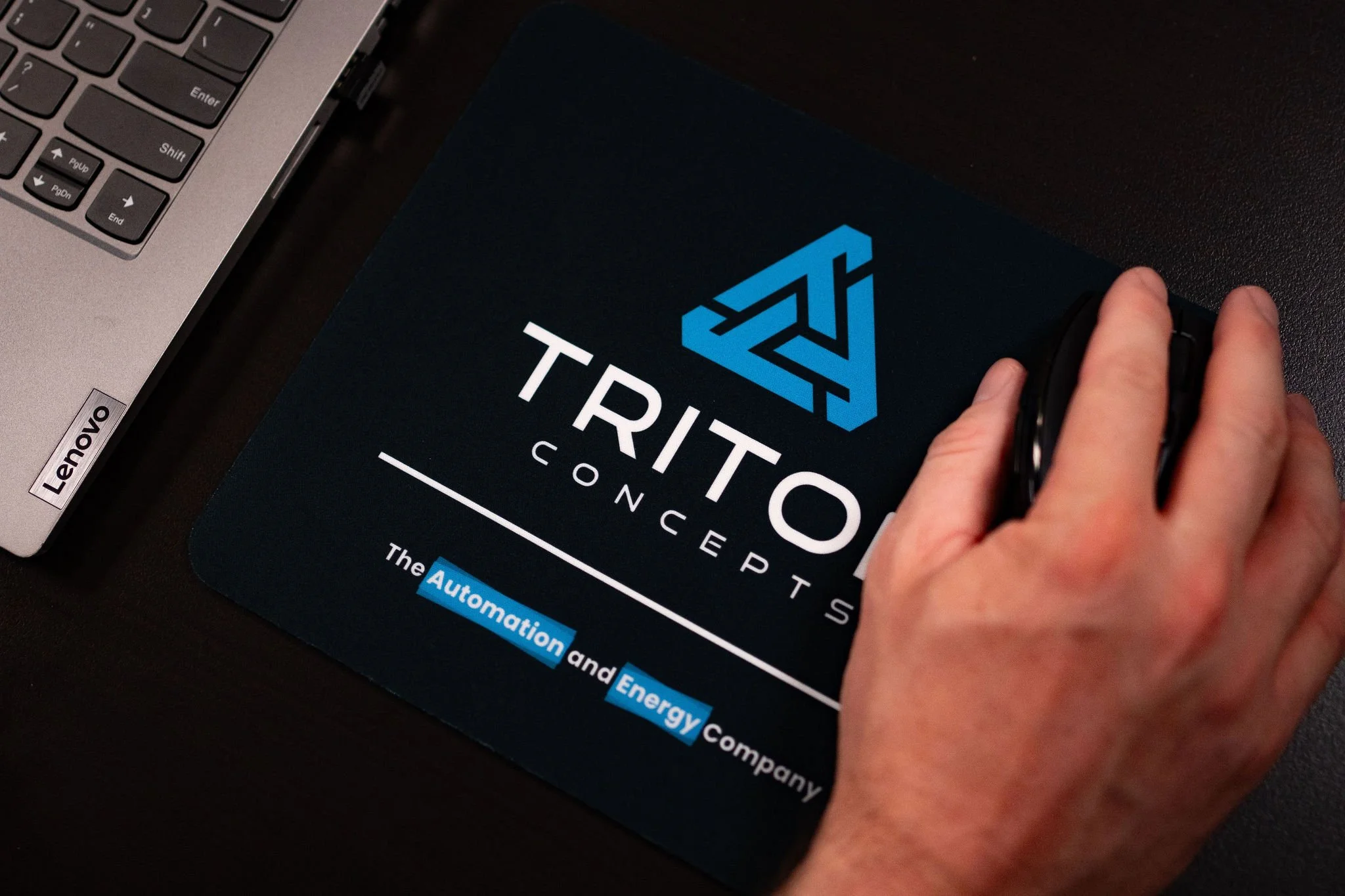 A hand resting on a computer mouse on a desk with a black mouse pad that has the Triton Concepts logo and text, next to a silver Lenovo laptop keyboard.
