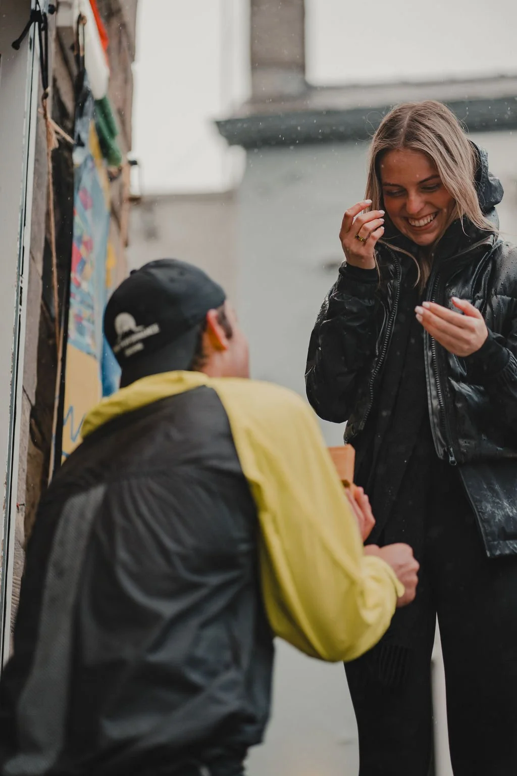 A woman smiling and laughing while talking to a person sitting on the ground outdoors in rainy weather. Both are dressed in jackets, and the woman has long hair.
