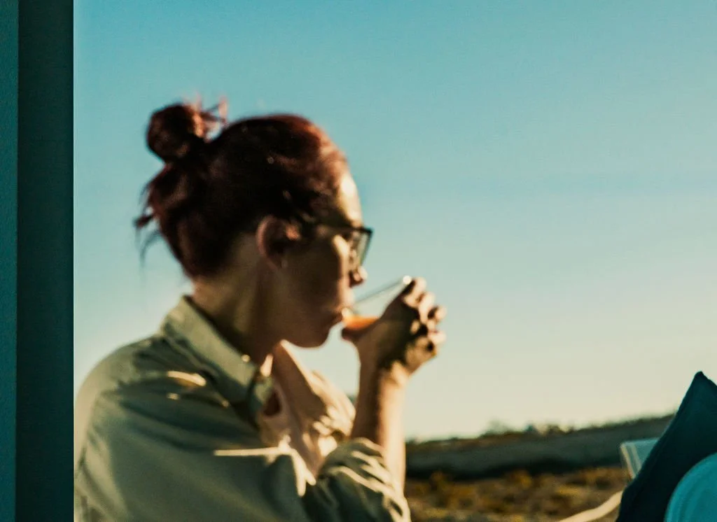 A woman with glasses and red hair tied in a bun drinking from a cup outdoors at sunset.