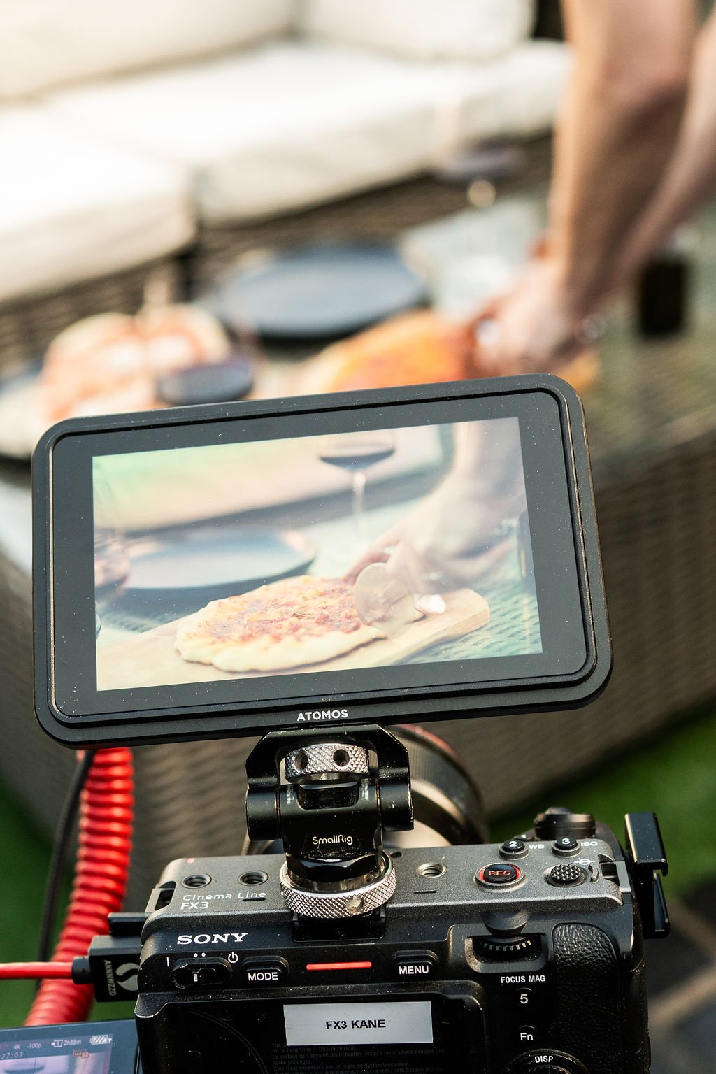 Camera monitor showing a person preparing a pizza on a wooden board outdoors, with additional cooking equipment in the background.