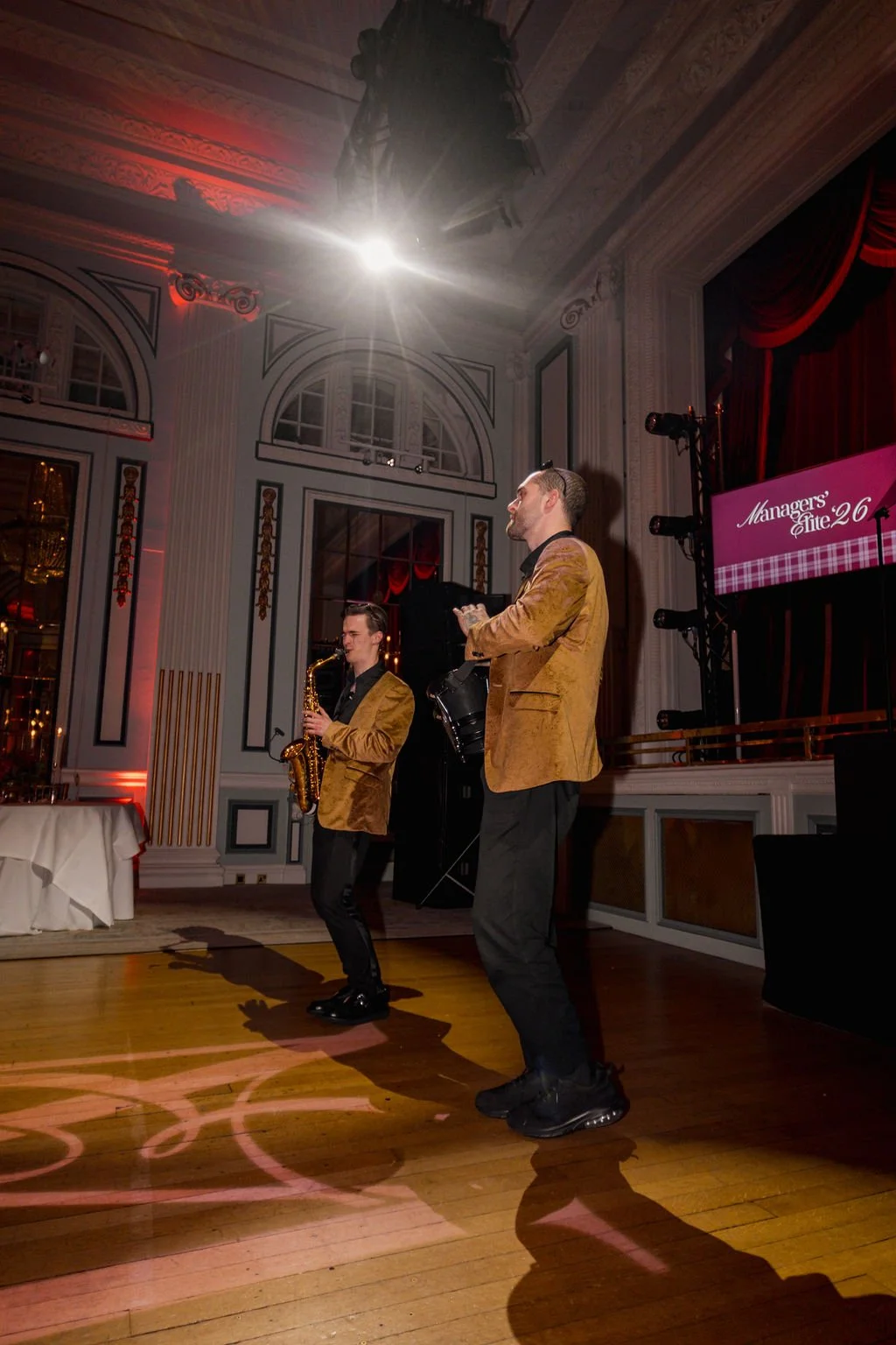 Two male performers in matching brown jackets dance and play saxophone and drums on stage in an ornate room with tall windows, decorative moldings, and a large chandelier. A large screen displays 'Managers' Elite 26'.