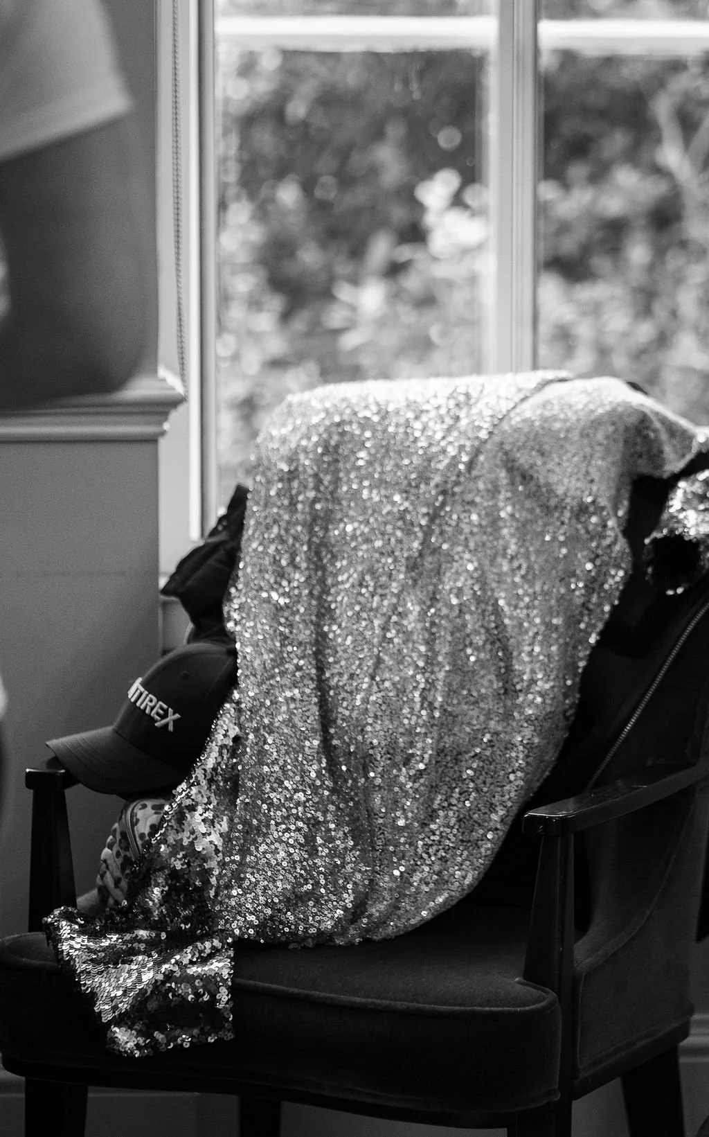 Sequin jacket draped over a chair with a baseball cap and a bag underneath, near a window with blurred outdoor scenery in the background.