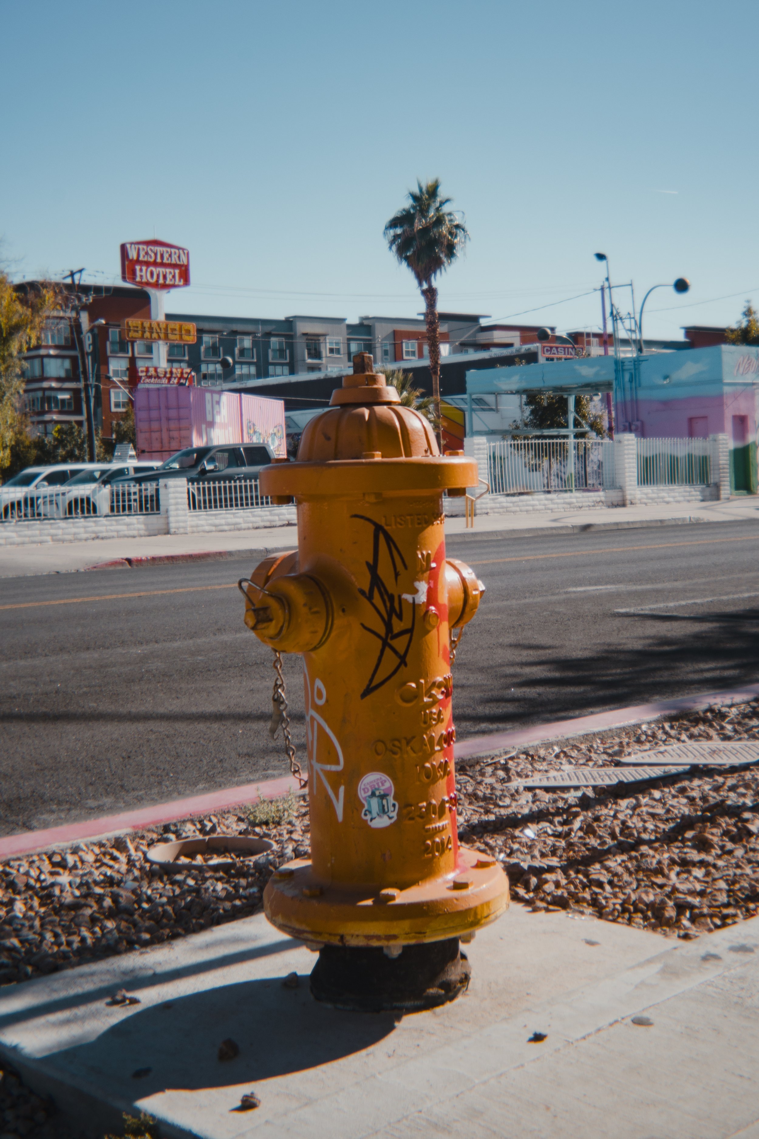 Orange fire hydrant with graffiti, located on a sidewalk next to a street with parked cars. In the background, there are colorful buildings, palm trees, and signs for a hotel and casino under a clear blue sky.