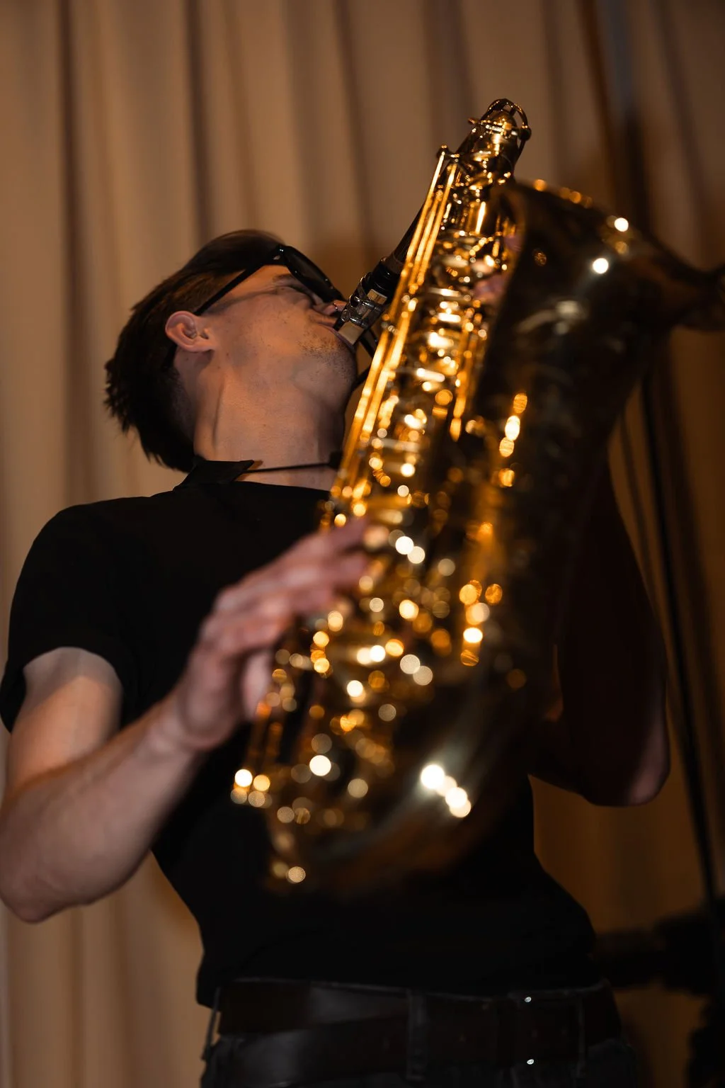 A man in a black T-shirt plays a gold saxophone, with his head tilted back and lips on the mouthpiece, in front of a beige curtain.