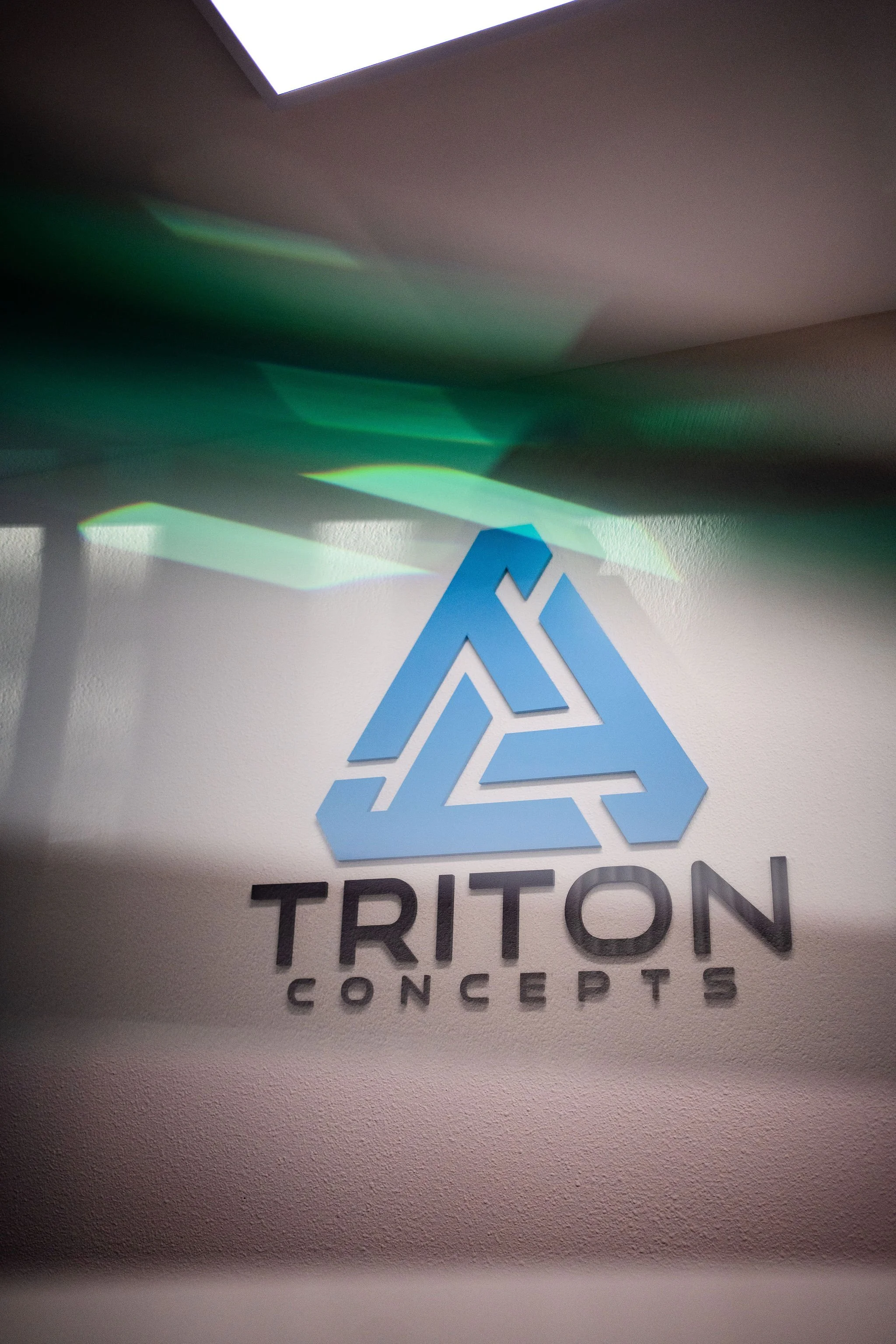 Close-up of a Triton Concepts logo on a wall, with a light overhead and some green and blue light reflections.