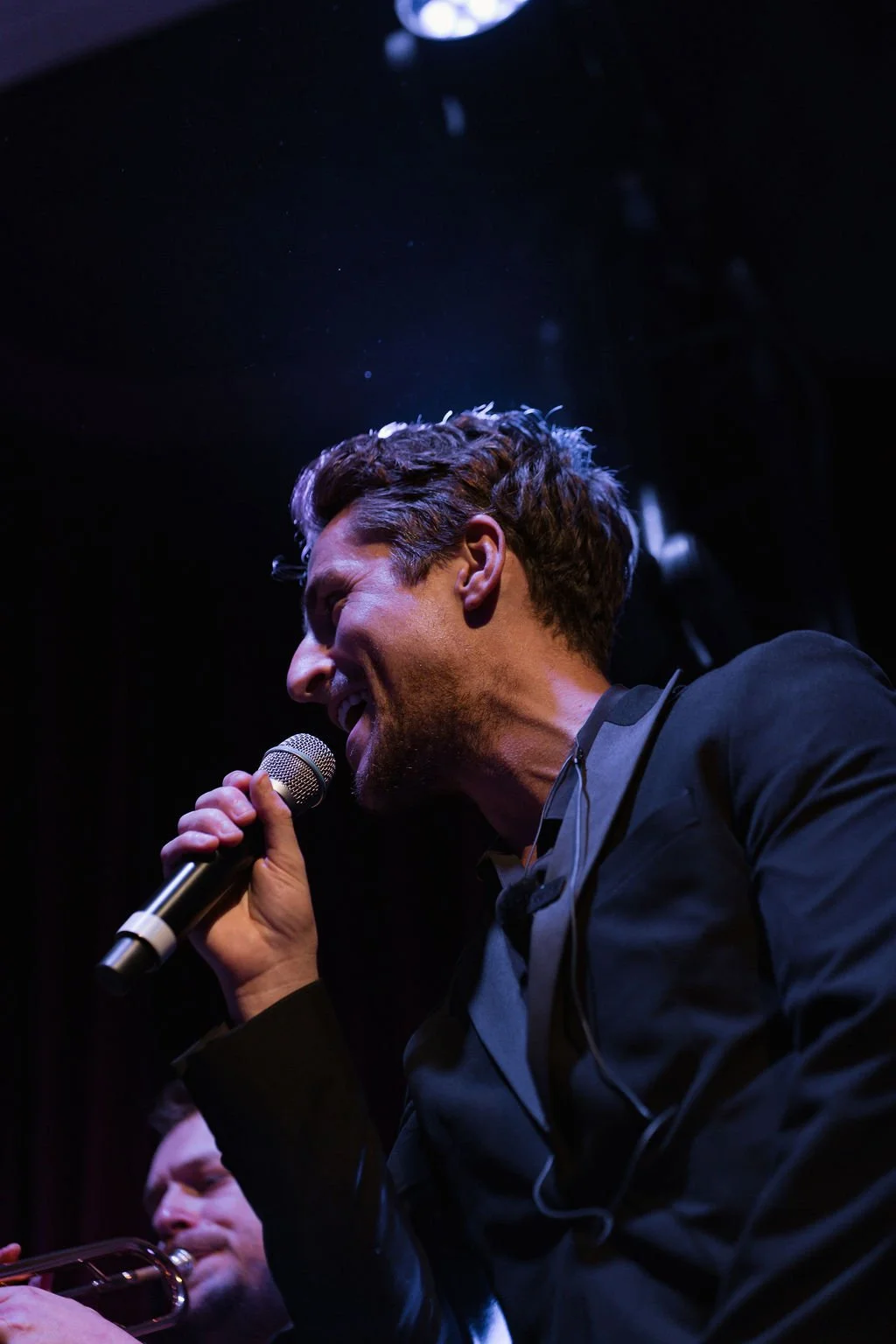 A man singing passionately into a microphone on stage with dark background and purple stage lighting.