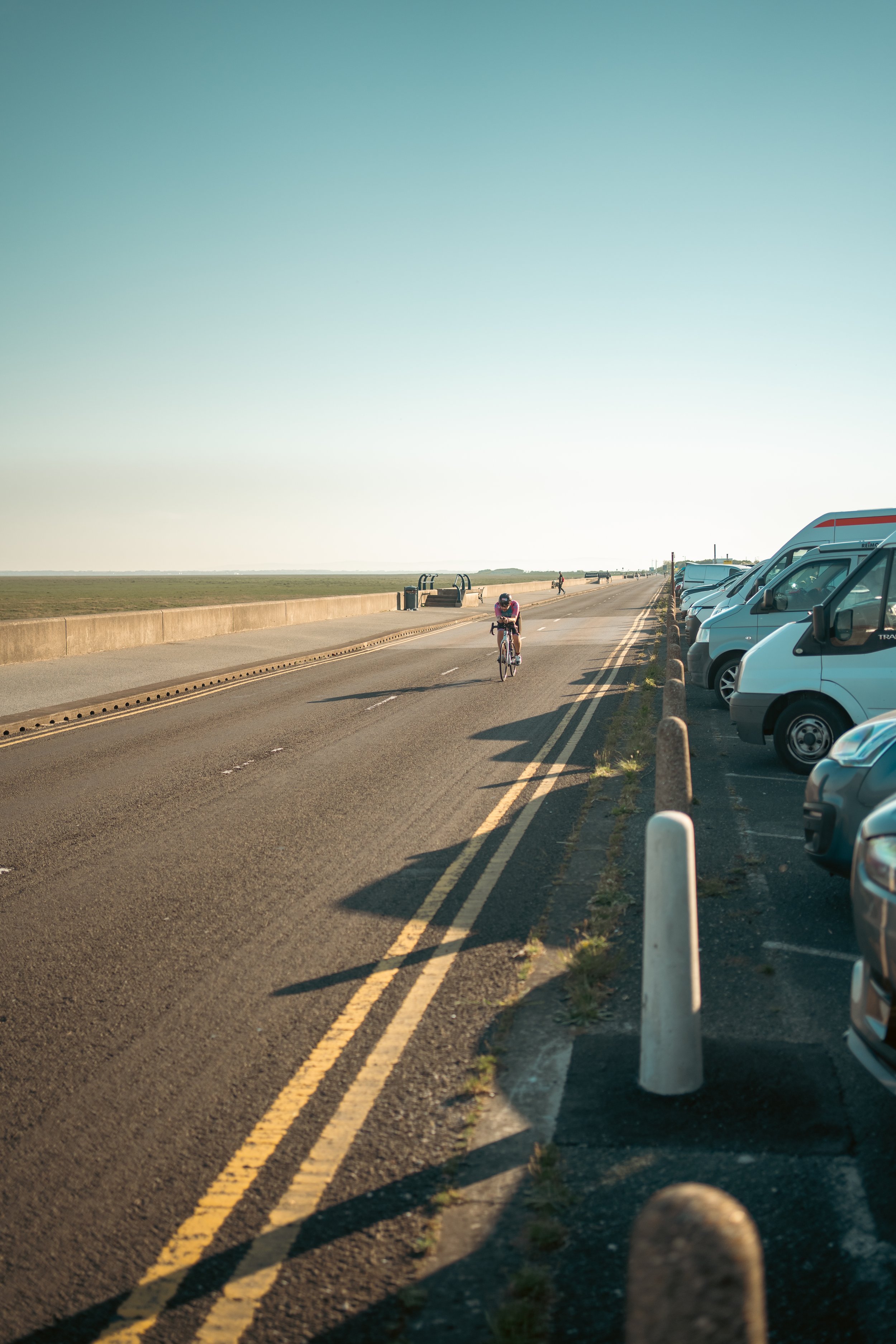 A cyclist riding along a coastal road with parked cars on the right side and an open grassy area on the left at sunset.