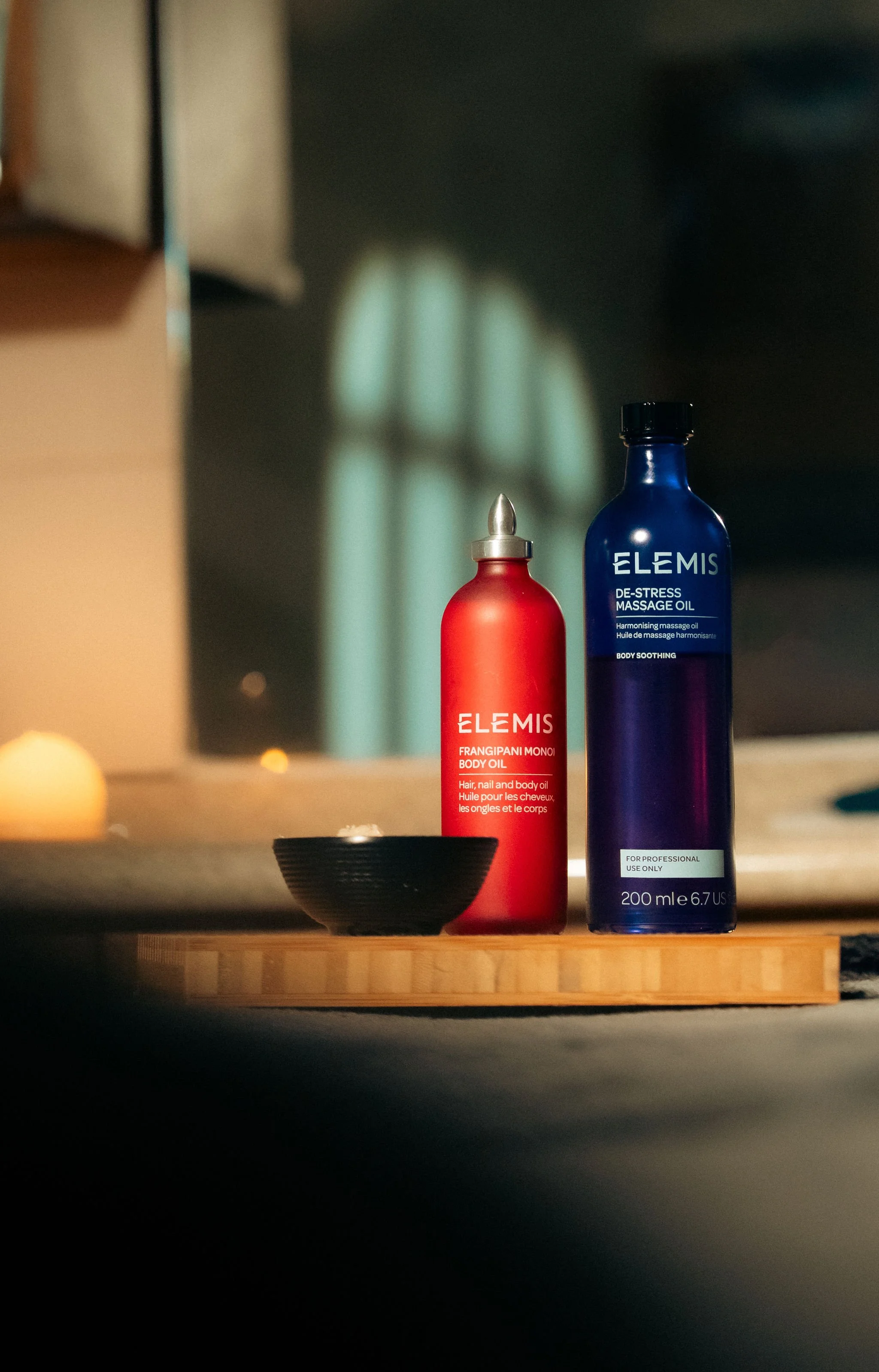 Two bottles of Elemis body oils, one red labeled 'Frangipani Monoi Body Oil' and one blue labeled 'De-Stress Massage Oil,' are placed on a wooden surface with a small black bowl nearby. Soft light and a window reflection are visible in the background