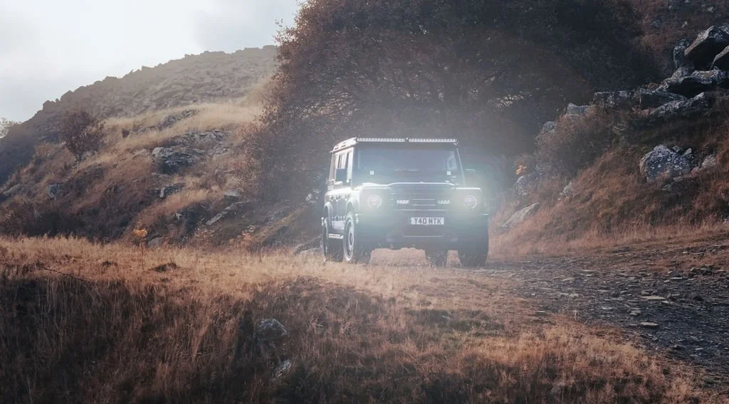 A dark green off-road vehicle driving on a rugged dirt trail through a hilly, grassy landscape with scattered rocks and bushes, and mountains in the background.