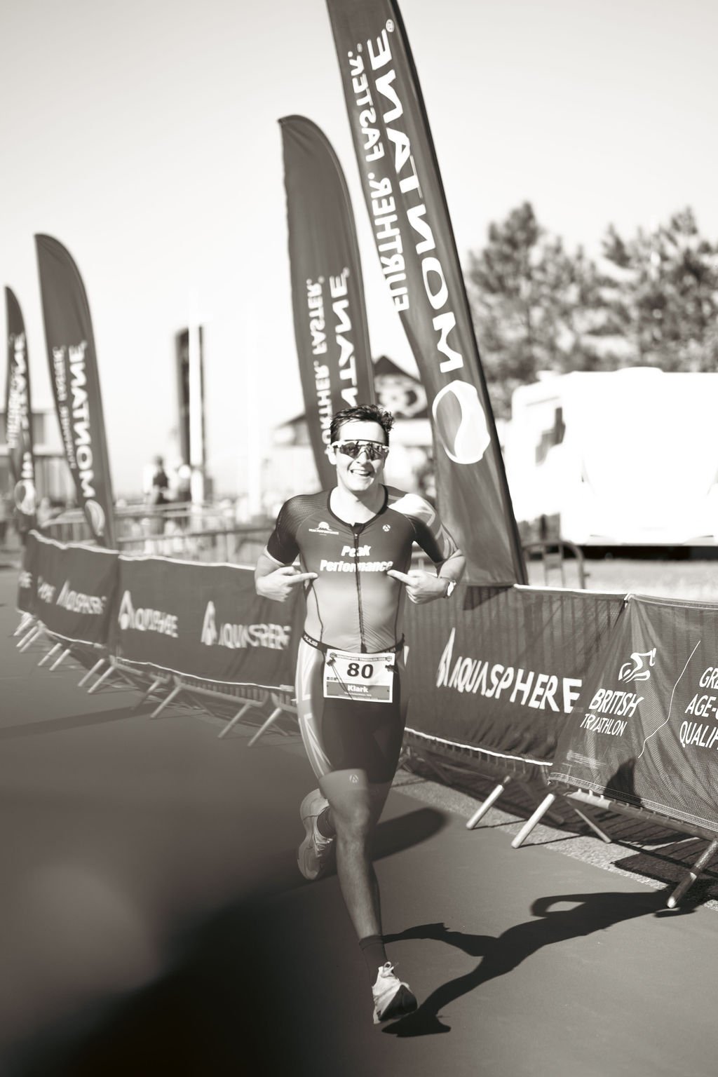 A male athlete running in a triathlon, smiling and pointing at himself, wearing sunglasses and a suit with the logo 'Peak Performance', numbered 80, at an outdoor event with banners and flags.