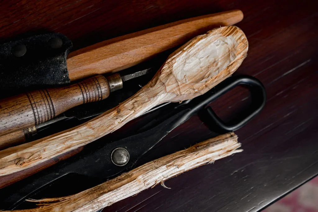 A collection of woodworking tools, including a wood-handled chisel, a black saw, and a wooden spoon, resting on a dark surface.