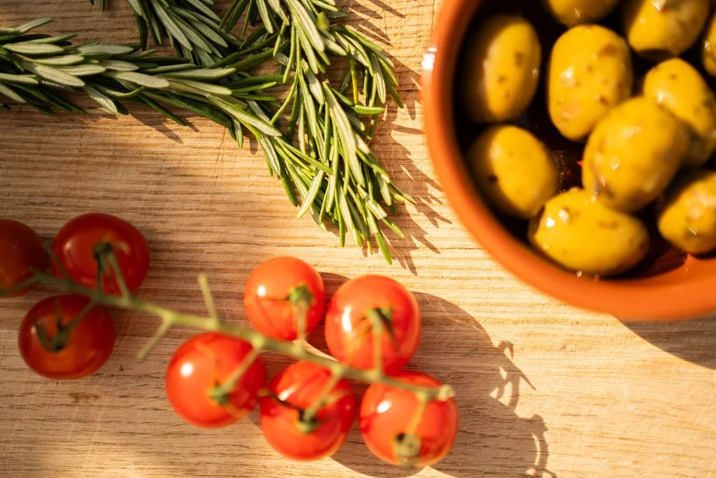 Fresh cherry tomatoes on vine, sprigs of rosemary, and a bowl of green olives on a wooden surface.
