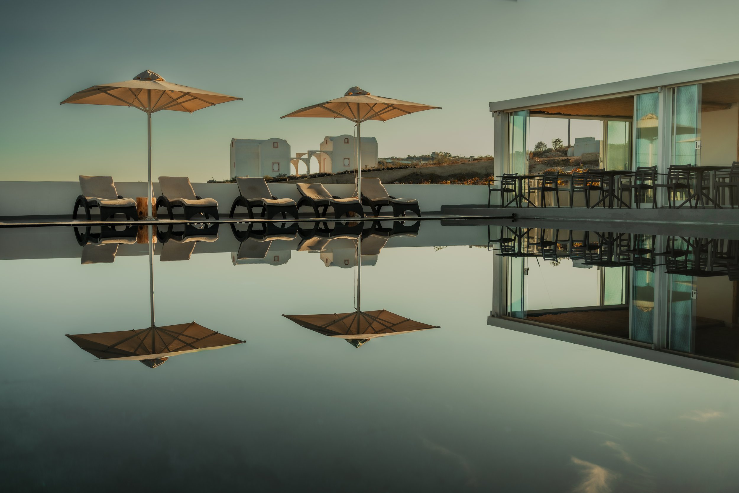 Outdoor pool area with lounge chairs and umbrellas, modern glass building, and scenic landscape in the background, reflected in the still water of the pool.