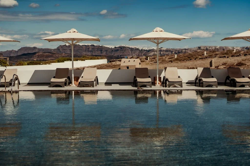 Empty poolside with lounge chairs, umbrellas, and a distant mountain landscape under a partly cloudy sky.