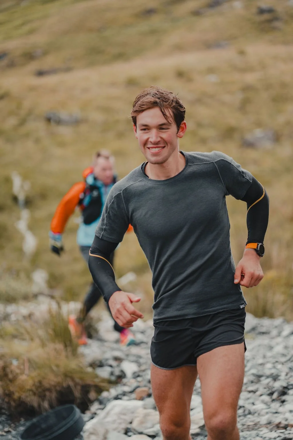 Young man running outdoors on rocky trail, smiling, with another person running behind in a grassy, mountainous landscape.