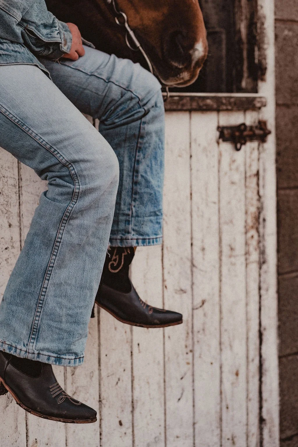 A person sitting on a wooden ledge with their legs dangling, wearing light blue jeans, black cowboy boots, and a denim jacket. There is a brown and white horse standing behind them with a bridle, and a rustic white wooden door with a latch in the bac