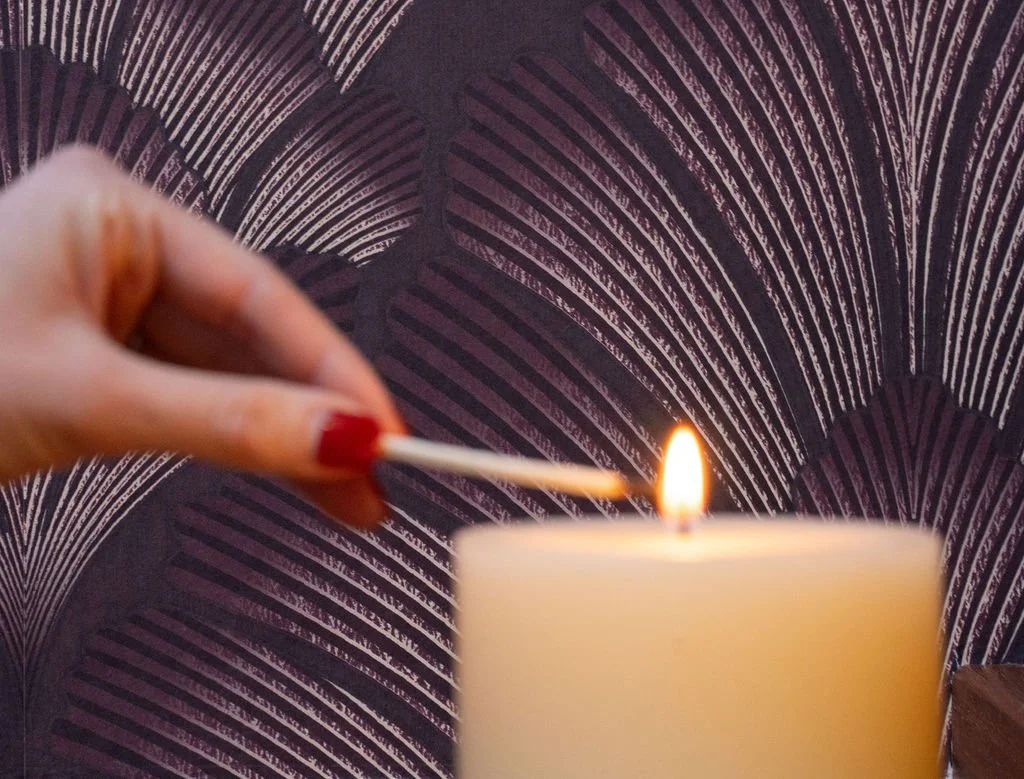 A person with red-painted fingernails lighting a candle with a matchstick in front of a patterned wallpaper background.
