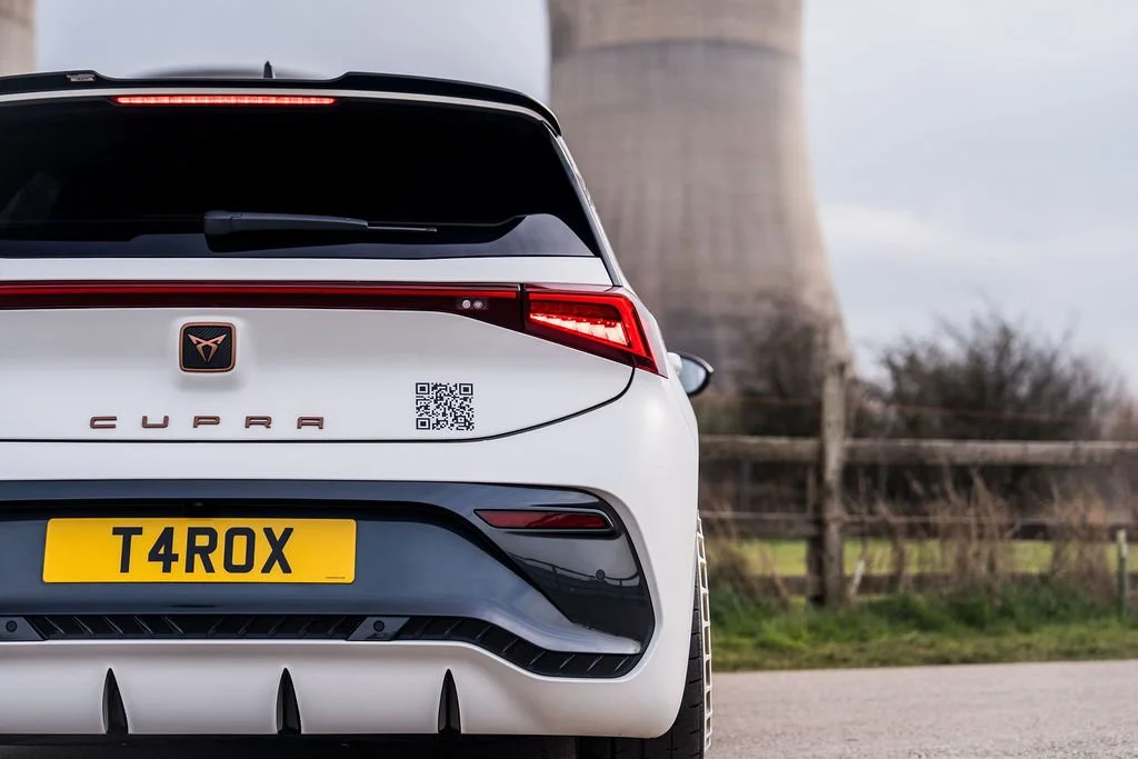 Rear view of a white Cupra T4ROX car with a black rear spoiler, QR code sticker, and yellow license plate, parked near a wooden fence and a power station.