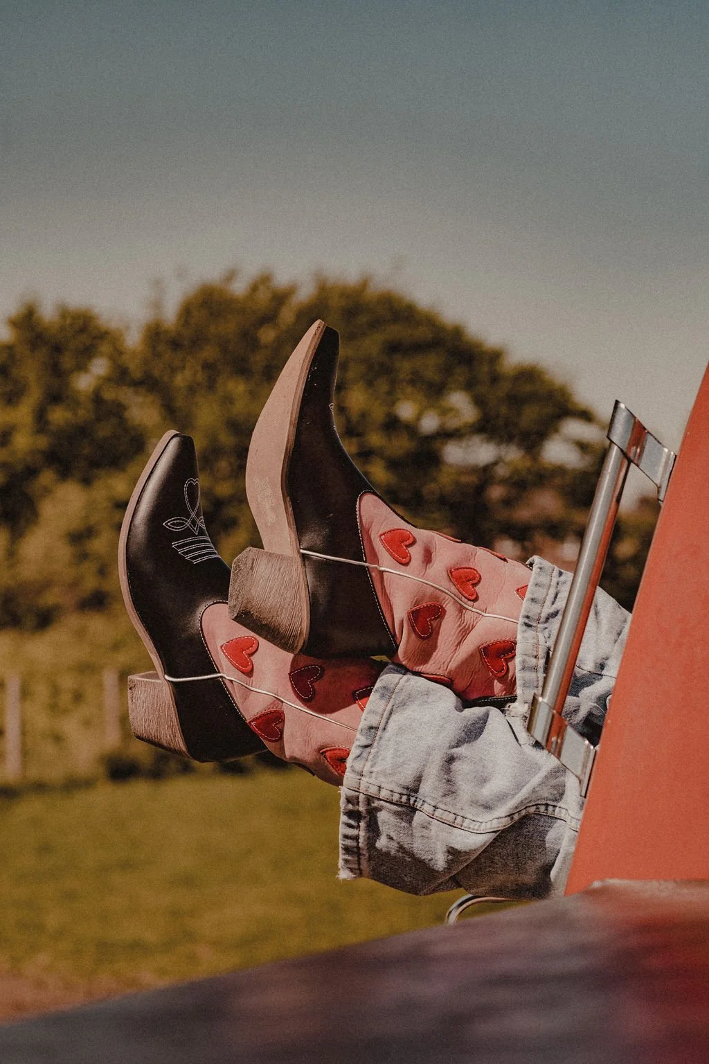 Person lying in truck with legs out of window, wearing cowboy boots with heart designs, blue jeans, and a pink skirt with red hearts. Trees and sky are blurred in the background.