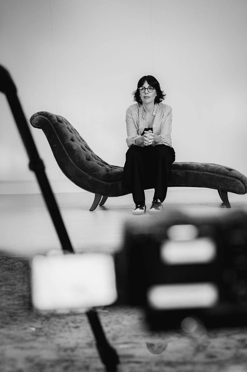 A woman with glasses and dark hair, wearing a striped shirt, sits on a vintage chaise lounge. The photo is in black and white, taken in a studio with a plain background.