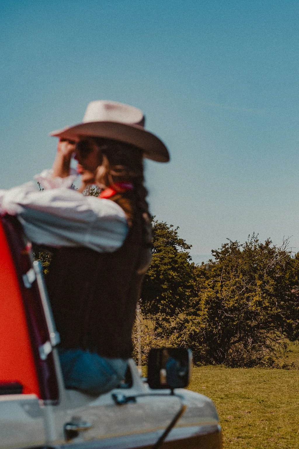 A woman wearing a cowboy hat and sunglasses, with braided hair, sitting in an off-road vehicle outdoors, with trees and a clear blue sky in the background.