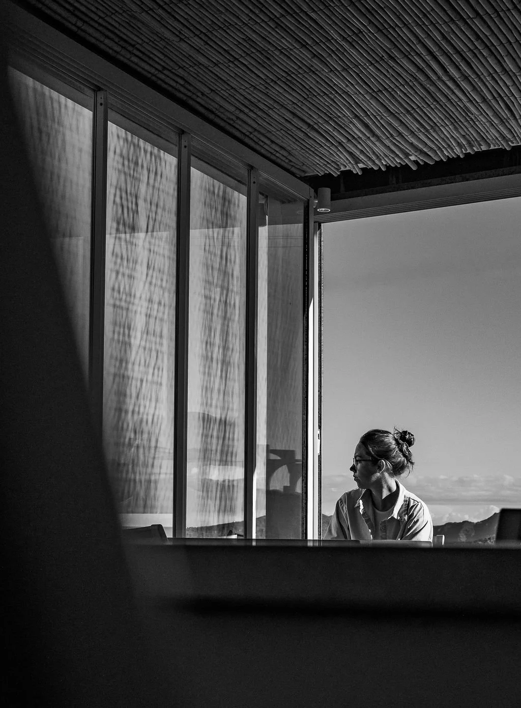 A woman with glasses sitting by a large window, looking to her right, with mountains in the background on a sunny day.