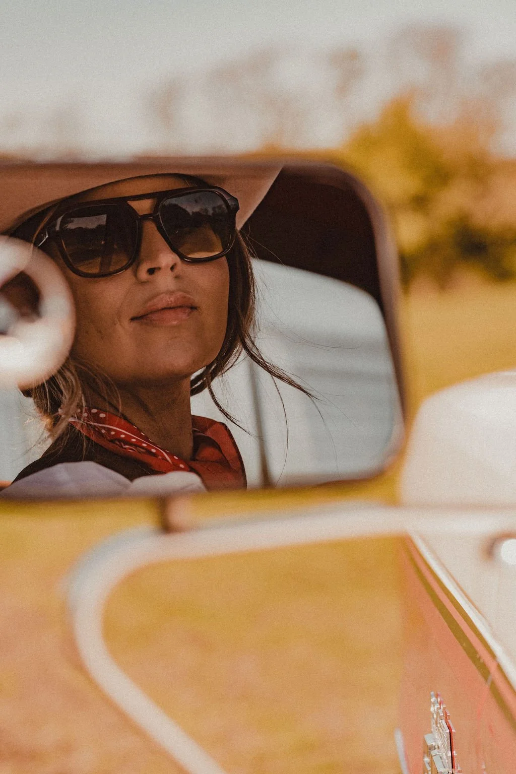 A woman wearing large sunglasses and a red bandana, looking out from a vehicle's side mirror.