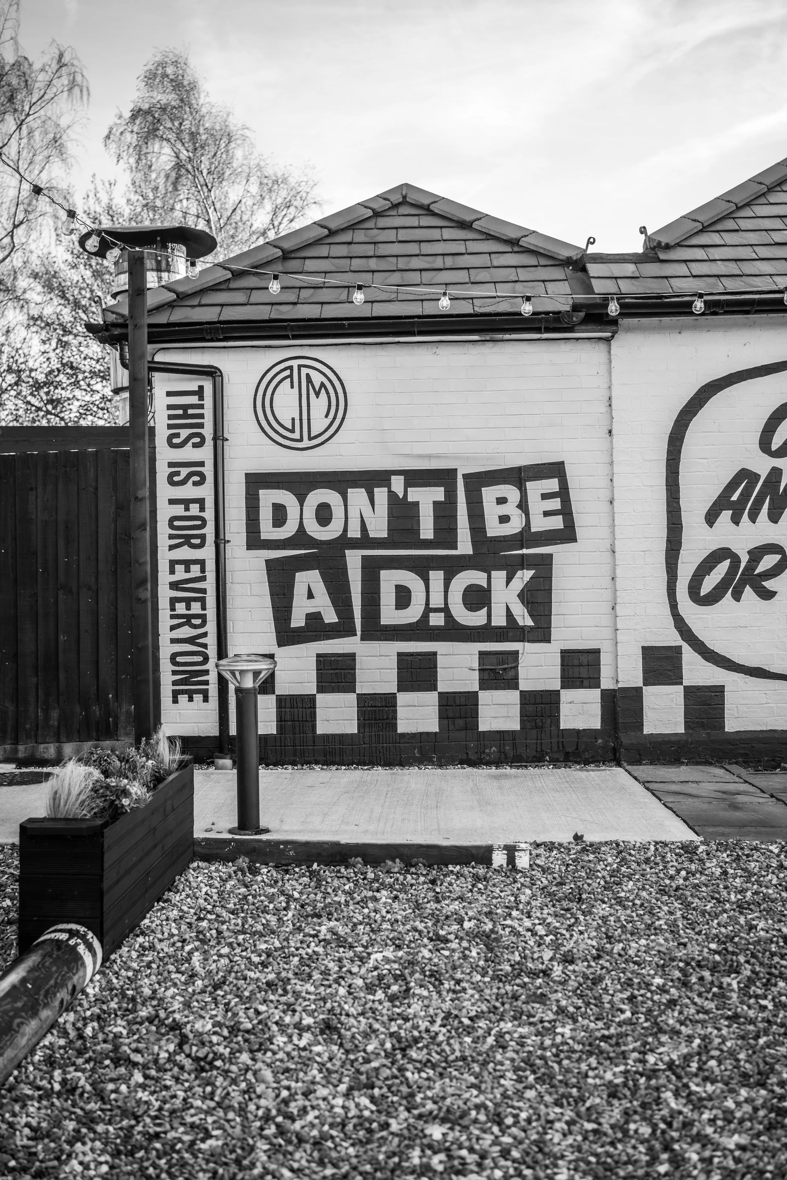 A black and white photo of a building with a mural that says, "DON’T BE A DICK," and a sign on the side that reads, "THIS IS FOR EVERYONE." There is outdoor lighting, a small gravel area, a planter with plants, and a sidewalk.