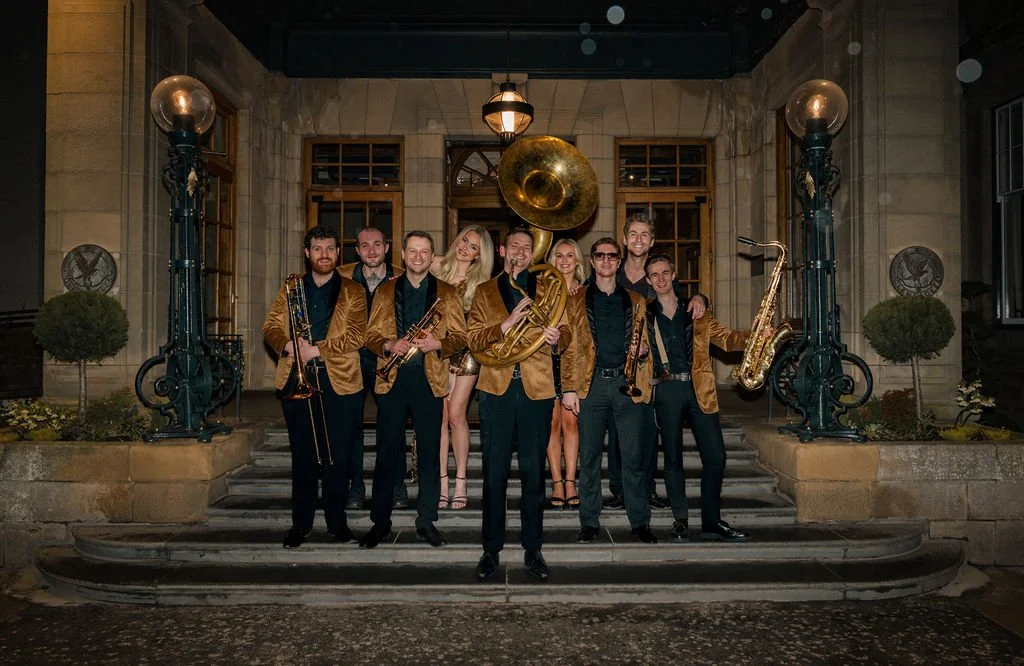 A group of nine musicians posing on the steps of a building at night, holding musical instruments including trumpets, a French horn, a saxophone, and a bassoon, with a large brass horn decoration hanging on the wall behind them.