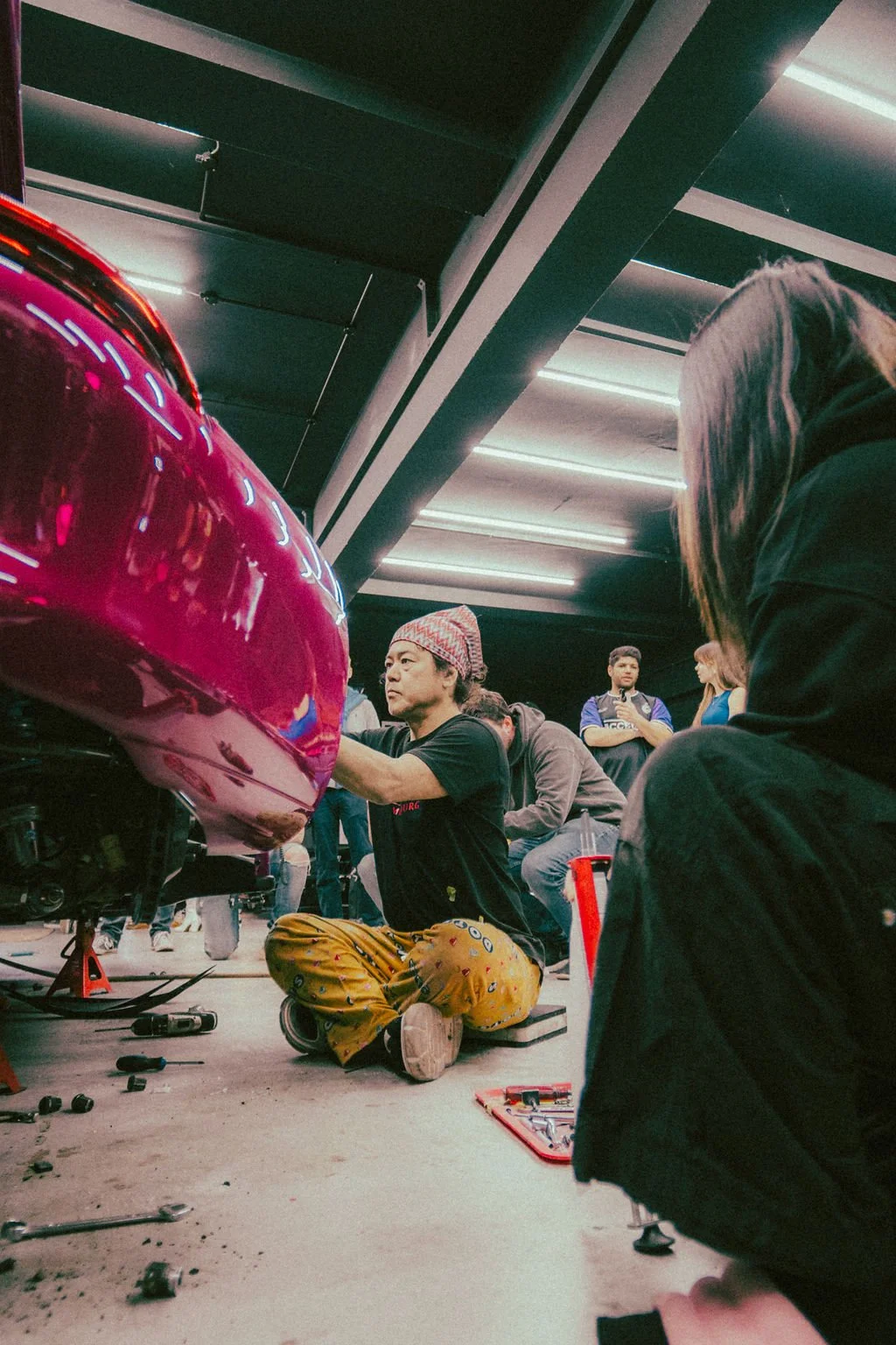 A man with a colorful beanie working on a car in a garage. Several people are observing and assisting, with tools and parts on the ground. Bright artificial lighting is overhead.